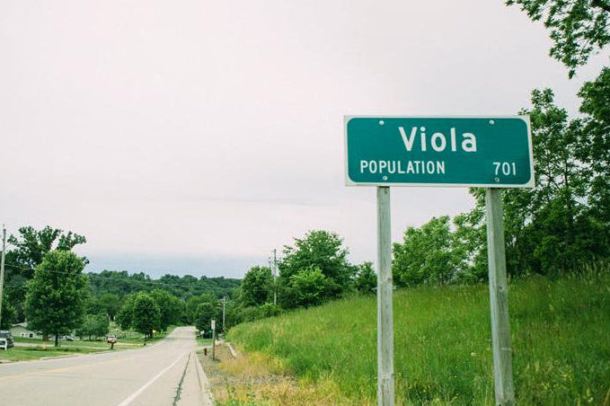 Street sign for Viola with a population of 701, set against a backdrop of trees and a road.